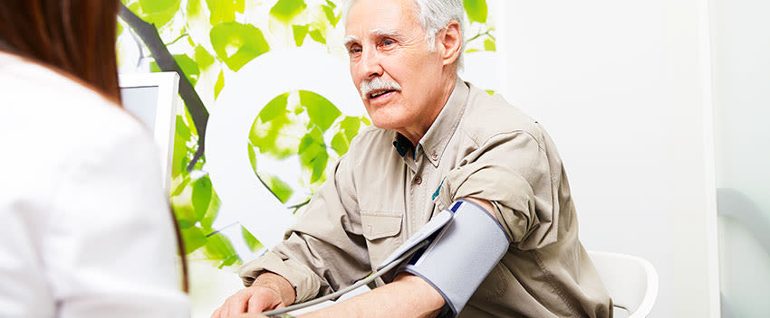 Man sitter hos en sjuksyster och får hjälp att ta blodtrycket med en blodtryckmätare.