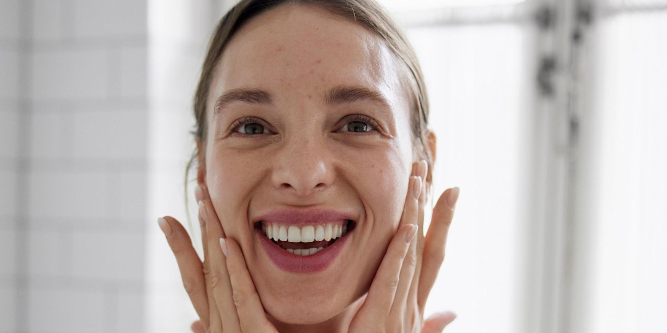 En kvinna med uppsatt hår ler stort och håller händerna vid ansiktet i en uttrycksfull och glad gest.