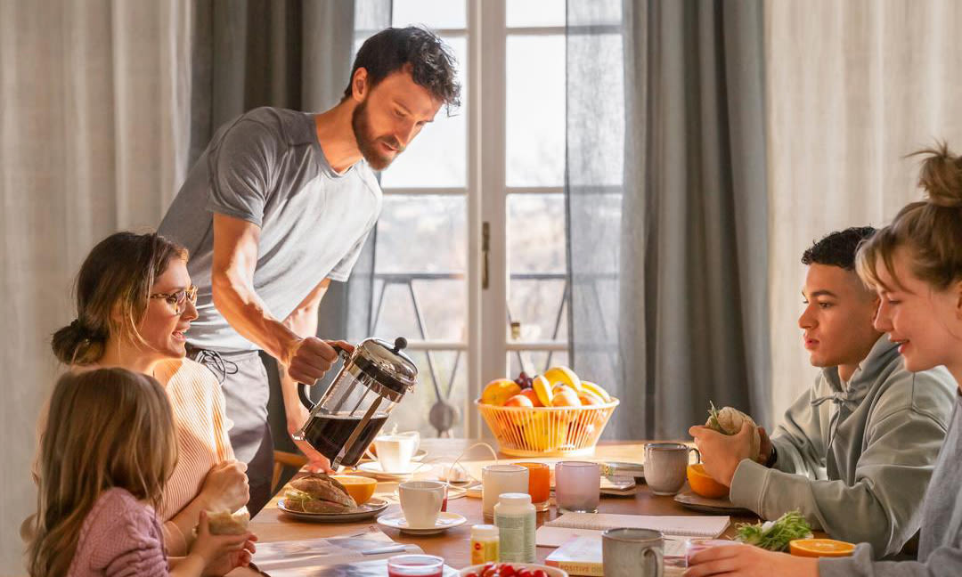Familj med en mamma, pappa, 2 tonåringar och ett yngre syskon sitter samlade vid frukostbordet. Bordet är dukat med en fruktskål med olika frukter i. Det står även kaffekoppar, glas med juice, bröd och olika pålägg. Pappan fyller på kaffe i en mugg medan de andra äter och dricker sin frukost.