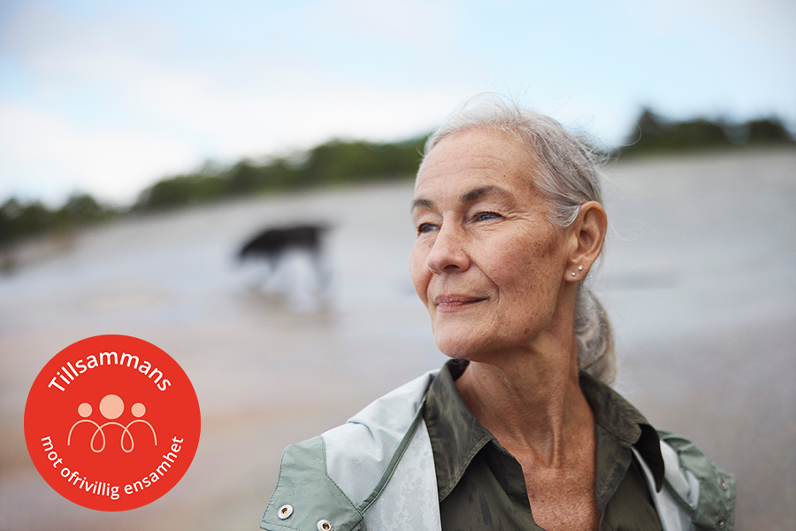 En medelålders kvinna med sin hund på stranden en regnig dag. En rödfärgad cirkel med den vita texten: "Tillsammans mot ofrivillig ensamhet" är synlig.